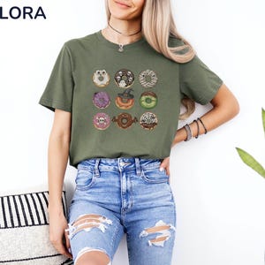 Puede incluir: Camiseta verde oliva con nueve donuts tem&aacute;ticos de Halloween. Cada donut tiene dise&ntilde;os &uacute;nicos, incluyendo una calavera, un sombrero de bruja y un murci&eacute;lago. La camiseta tiene el texto "MELORA SHIRTS" en la parte superior izquierda.