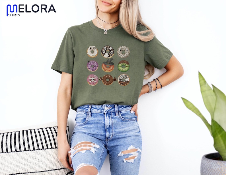 Puede incluir: Camiseta verde oliva con nueve donuts tem&aacute;ticos de Halloween. Cada donut tiene dise&ntilde;os &uacute;nicos, incluyendo una calavera, un sombrero de bruja y un murci&eacute;lago. La camiseta tiene el texto "MELORA SHIRTS" en la parte superior izquierda.