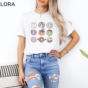 Puede incluir: Camiseta blanca con un gr&aacute;fico de nueve donuts tem&aacute;ticos de Halloween. Cada donut est&aacute; decorado con dise&ntilde;os espeluznantes como calaveras y sombreros de bruja. La camiseta tiene la marca "MELORA SHIRTS" en la esquina superior izquierda.
