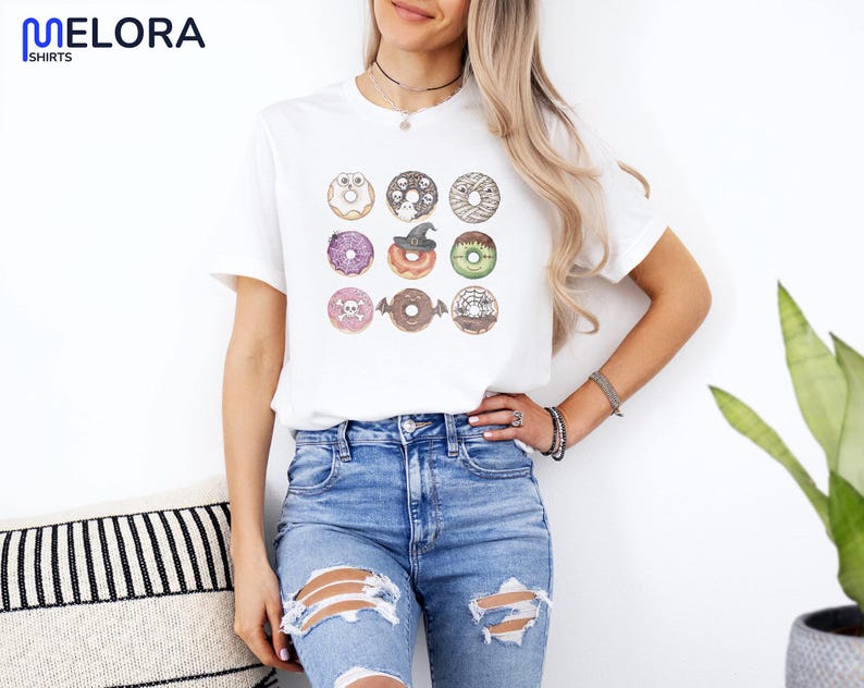 Puede incluir: Camiseta blanca con un gr&aacute;fico de nueve donuts tem&aacute;ticos de Halloween. Cada donut est&aacute; decorado con dise&ntilde;os espeluznantes como calaveras y sombreros de bruja. La camiseta tiene la marca "MELORA SHIRTS" en la esquina superior izquierda.