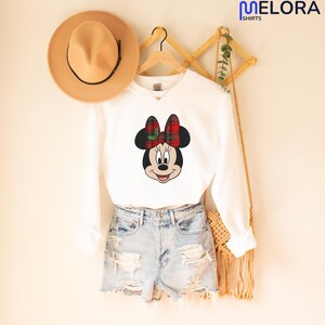 May include: A white long-sleeved sweatshirt with a Minnie Mouse design, sporting a red and green tartan bow. The outfit includes denim shorts and a tan hat. The Melora Shirts logo is visible in the top right corner.