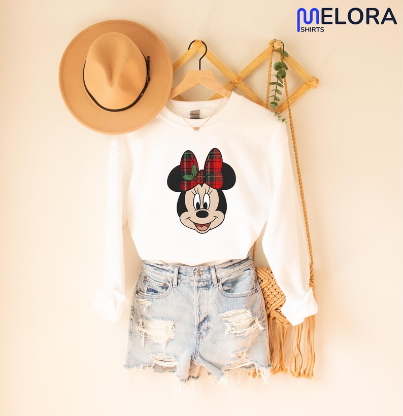 May include: A white long-sleeved sweatshirt with a Minnie Mouse design, sporting a red and green tartan bow. The outfit includes denim shorts and a tan hat. The Melora Shirts logo is visible in the top right corner.