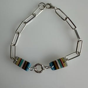 Può includere: Bracciale a catena argentato con due perline rettangolari a righe blu, marroni e gialle. Il bracciale ha una chiusura a moschettone e un piccolo connettore rotondo.