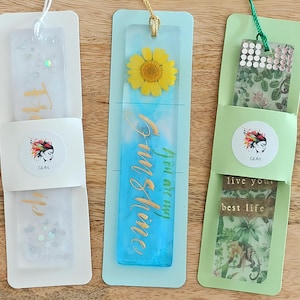 May include: Three handmade bookmarks with different designs. The first bookmark is white with glitter and the text "Believe" written vertically. The second bookmark is blue with a pressed flower and the text "You are my sunshine". The third bookmark is green with a pressed leaf design and the text "Live your best life".