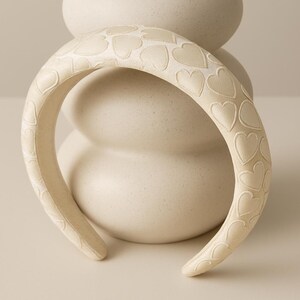 May include: Cream-colored headband with an embossed heart pattern. The headband is positioned in front of a white, textured vase with brown stems. The design is simple and elegant, suitable for various occasions.