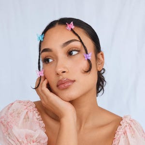 May include: A set of four small, translucent butterfly hair clips in shades of pink, blue, and purple, attached to two thin, braided strands of dark hair. The clips feature a glossy finish and are positioned along the hair strands.