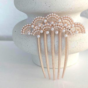 May include: A rose gold hair comb featuring a decorative fan-shaped top adorned with small white faux pearls. The comb has five sturdy, textured prongs designed for securing hair, with the pearl embellishments arranged in a scalloped pattern.