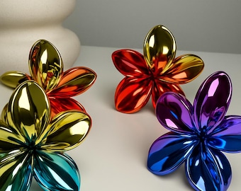 Könnte beinhalten: Fünf metallische, blütenförmige Dekorationsgegenstände in leuchtenden Farben: Gold, Rot, Orange, Blau und Lila. Jede Blume hat fünf Blütenblätter und eine reflektierende, glänzende Oberfläche. Die Objekte sind auf einer weißen Oberfläche angeordnet.
