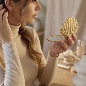 Op de afbeelding: Een goudkleurige, schelpvormige compacte spiegel, open om een reflecterend oppervlak te onthullen. De compact is in een hand gehouden, met een bijpassende gesloten compact op een houten oppervlak. De algehele esthetiek is elegant en decoratief.