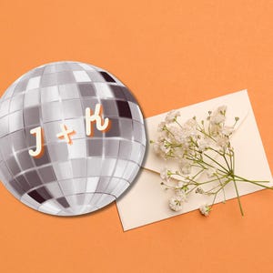 May include: A silver disco ball with the letters "J + K" in orange on a light orange background. A white envelope with a sprig of white flowers is in the lower right corner.