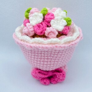 May include: A crocheted bouquet of pink, white, and green flowers in a pink crocheted vase with a pink bow.