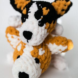 May include: A crocheted stuffed toy dog with brown, white, and black fur. The dog is sitting up with its head tilted to the side.