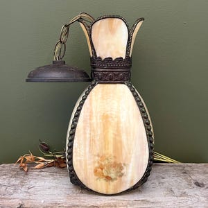 May include: A hanging pendant lamp featuring a teardrop-shaped, cream-coloured marbled glass shade with a floral decal. The lamp is encased in a dark bronze-toned metal frame with decorative beaded edges and a matching circular ceiling canopy.