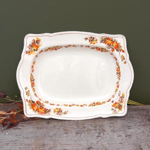 Peut inclure: Un plat de service rectangulaire en céramique avec des bords festonnés et une base blanche. Le bord présente une bordure décorative de feuilles d'érable aux couleurs automnales orange, jaune et marron, avec un fin liseré doré sur le pourtour.