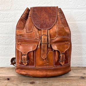 Puede incluir: Una mochila de cuero marrón con cierre de solapa y dos bolsillos delanteros. La mochila presenta costuras blancas y hebillas color latón. El cuero tiene un aspecto envejecido, y la bolsa está colocada contra una pared de ladrillos blancos y una superficie de madera.