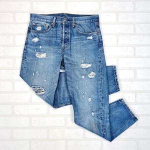 Op de afbeelding: Lichtblauwe denim jeans met distressed details, waaronder scheuren en vervaagde vlekken. De jeans hebben een knoopsluiting en een klassiek ontwerp met vijf zakken. Het kledingstuk ligt plat tegen een witte bakstenen muur.