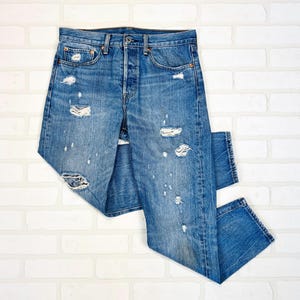 Vaqueros Levi's 501 azules desgastados con rotos y lavados pronunciados, corte recto clásico, cintura alta y cierre de botones.