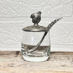 May include: A clear glass jar with a silver-toned metal lid and a matching silver-toned metal spoon. The lid features a sculpted animal figure and intricate engraved patterns. The spoon handle also displays detailed engraved markings.
