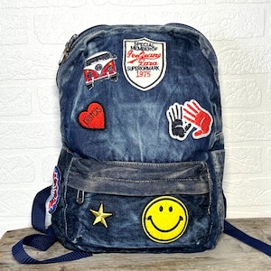 Puede incluir: Mochila vaquera azul con varios parches. Los parches incluyen un corazón rojo con texto, una carita sonriente amarilla, una estrella dorada y un autobús VW rojo y blanco. La mochila tiene una correa azul para el hombro y cierre de cremallera.