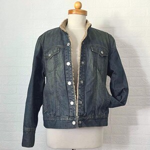 May include: A dark wash denim jacket with a tan sherpa lining and collar. The jacket features two chest pockets, two side pockets, and white button fastenings. The jacket is displayed on a mannequin.