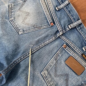 May include: Close-up of light blue denim jeans with visible pockets and stitching. The Wrangler brand label is visible on a brown leather patch. The jeans show signs of wear and tear.