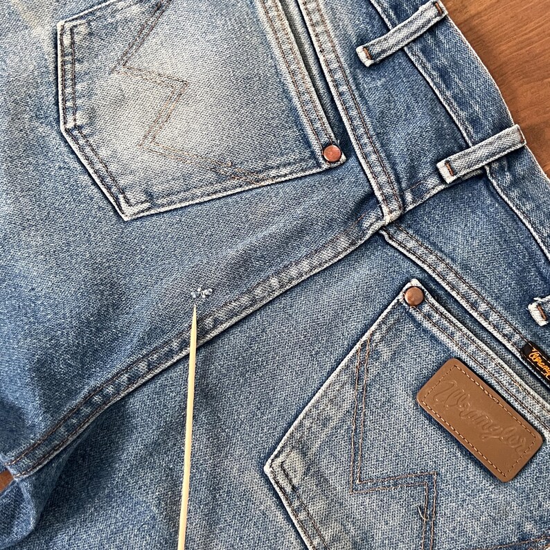 May include: Close-up of light blue denim jeans with visible pockets and stitching. The Wrangler brand label is visible on a brown leather patch. The jeans show signs of wear and tear.