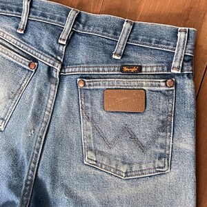 May include: Close-up of the back of light blue denim jeans. The jeans feature a brown leather patch with the "Wrangler" logo on the back pocket. The jeans have copper rivets and a small "Wrangler" label above the back pocket.