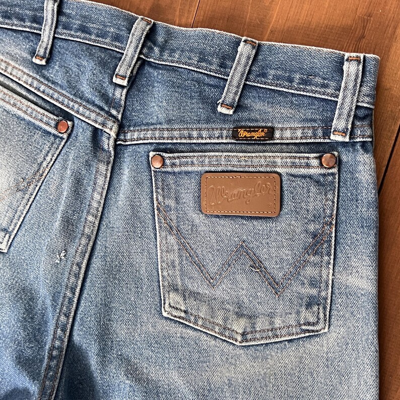 May include: Close-up of the back of light blue denim jeans. The jeans feature a brown leather patch with the "Wrangler" logo on the back pocket. The jeans have copper rivets and a small "Wrangler" label above the back pocket.