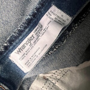 May include: Close-up of a pair of blue denim Wrangler jeans. The white tag displays the brand name, size, and care instructions. The jeans are made of 100% cotton and manufactured in Nicaragua. The tag also includes the style number, 13MWZ, and the size, 31 x 34.