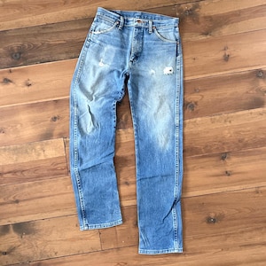 May include: Light blue denim jeans with a classic straight leg fit. The jeans show signs of wear with distressed areas on the front. Features include a button and zipper closure, front and back pockets, and belt loops. The jeans are laid flat on a wooden surface.