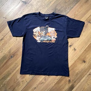 Puede incluir: Camiseta azul marino con un dibujo animado de un coche y el texto "Midnight Hustler". El gráfico incluye elementos naranjas, blancos y negros. La camiseta está hecha de un material suave.