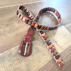 Op de afbeelding: Een gevlochten leren riem met een rechthoekige gesp. De riem heeft een patroon van bruine, oranje en groene leren stukken. De gesp en de riemlus zijn roodbruin van kleur.