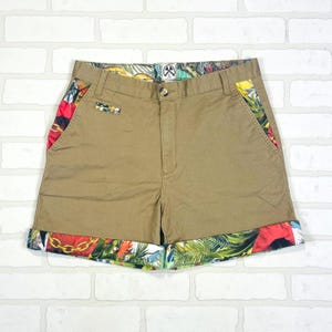 Puede incluir: Pantalones cortos color caqui con un estampado tropical colorido en los bolsillos y los puños. Los pantalones cortos tienen cierre de botón y un pequeño bolsillo en la parte delantera. El estampado tropical incluye colores rojo, amarillo y verde.