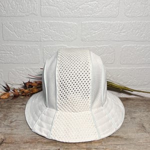 May include: A white bucket hat featuring a structured design with alternating panels of solid fabric and breathable mesh. The hat has a wide, downward-sloping brim and a rounded crown, constructed from lightweight, textured material in a uniform white colour.