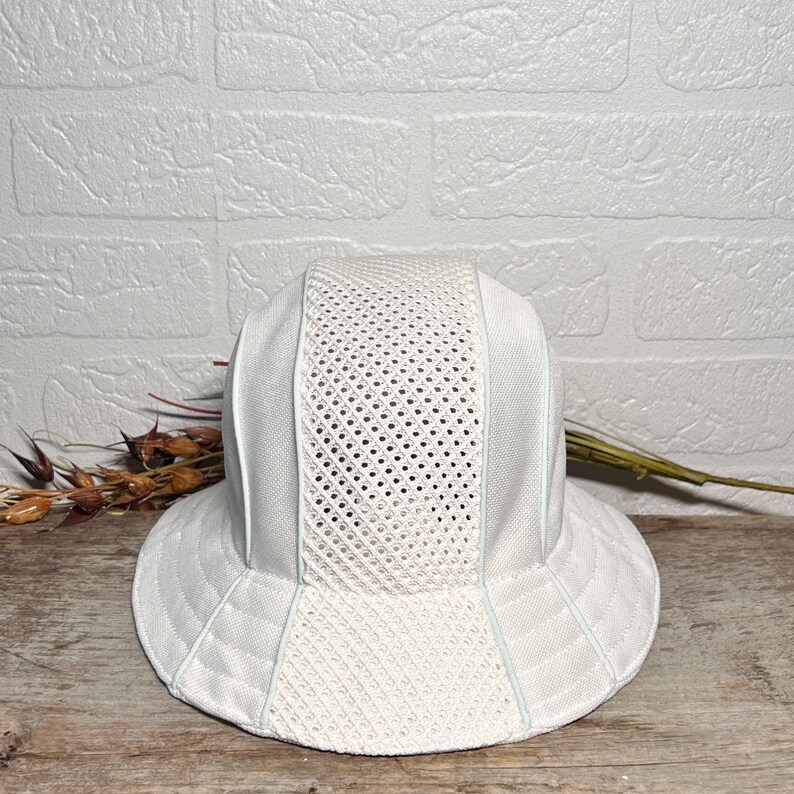 May include: A white bucket hat featuring a structured design with alternating panels of solid fabric and breathable mesh. The hat has a wide, downward-sloping brim and a rounded crown, constructed from lightweight, textured material in a uniform white colour.