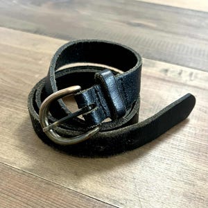 以下が含まれることがあります： シルバー色のバックルが付いた黒い革ベルト。ベルトは木製の表面に巻かれており、その質感とデザインを示しています。バックルはクラシックでシンプルなスタイルです。