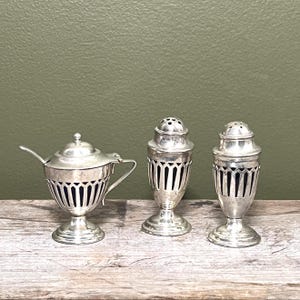 May include: A silver-coloured condiment set comprising a lidded mustard pot with a small spoon and two matching shakers. Each piece features a pedestal base and a decorative openwork cage design that reveals a dark blue glass insert inside.
