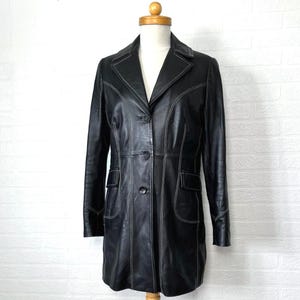 May include: A black leather jacket with white stitching. The jacket has a long, fitted silhouette and a classic collar. It features a button closure and two front pockets.