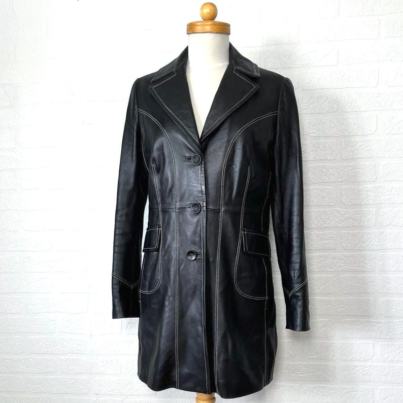 May include: A black leather jacket with white stitching. The jacket has a long, fitted silhouette and a classic collar. It features a button closure and two front pockets.