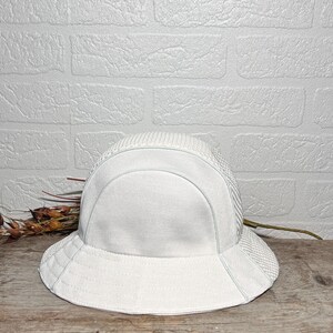 May include: A solid white bucket hat featuring a structured crown and a wide, downward-sloping brim. The hat is constructed from a textured, woven fabric with a distinct mesh-like pattern on the side panels and a smooth, solid finish on the front.