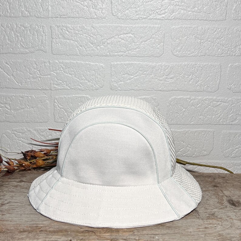 May include: A solid white bucket hat featuring a structured crown and a wide, downward-sloping brim. The hat is constructed from a textured, woven fabric with a distinct mesh-like pattern on the side panels and a smooth, solid finish on the front.