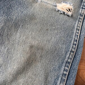 May include: Close-up of light blue denim jeans with distressed details. The jeans have a ripped section revealing frayed threads. The fabric shows subtle variations in color, with a visible seam on the right side.