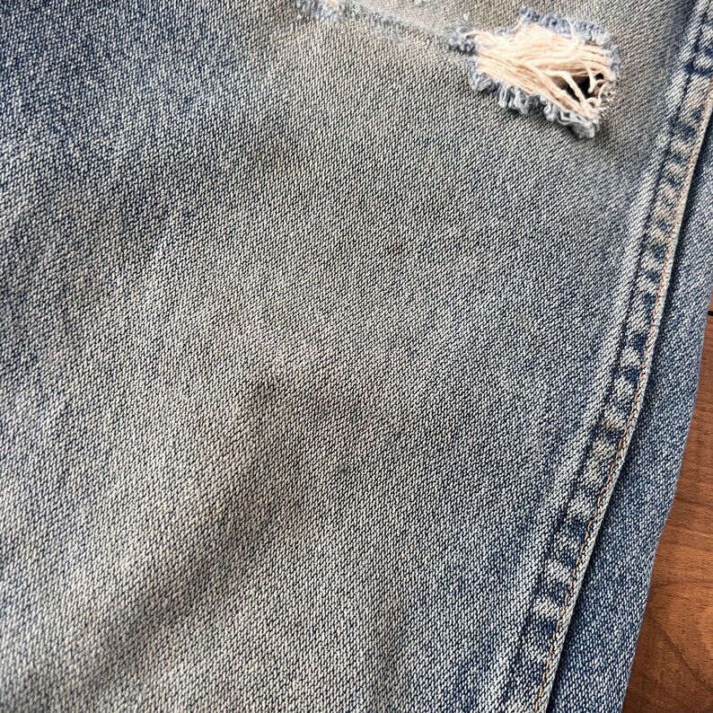 May include: Close-up of light blue denim jeans with distressed details. The jeans have a ripped section revealing frayed threads. The fabric shows subtle variations in color, with a visible seam on the right side.