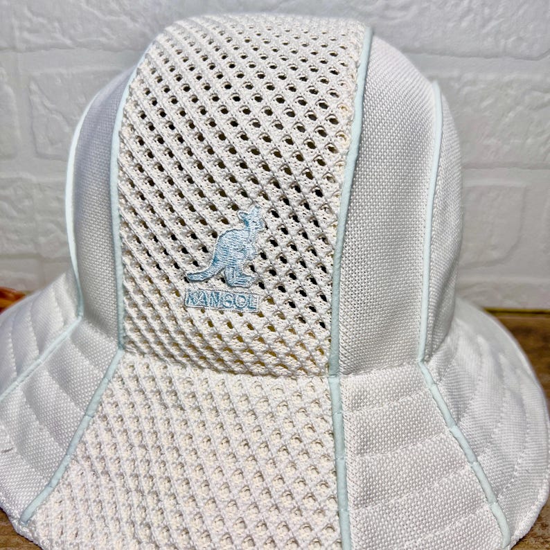 May include: A white bucket hat featuring a central mesh panel and solid side panels. The front of the hat displays an embroidered light blue Kangol kangaroo logo with the text KANGOL underneath. The hat has a wide brim and light blue piping.