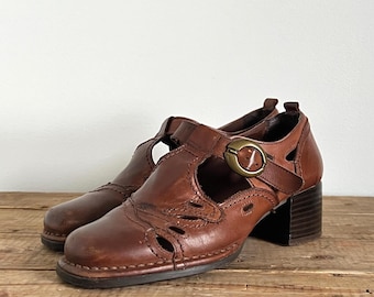 May include: A pair of brown leather shoes featuring a T-strap design, cut-out details along the vamp, and a rounded toe. The shoes include a side strap with a circular brass-toned buckle and a stacked block heel with a dark brown finish.