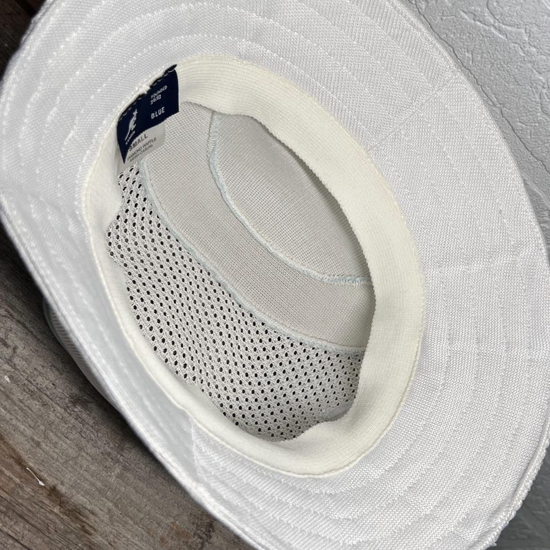 May include: A white bucket hat viewed from the inside, featuring a mesh lining for ventilation and a sweatband along the inner rim. A small dark tag is attached to the interior, displaying the Kangol logo and the size label marked as small.