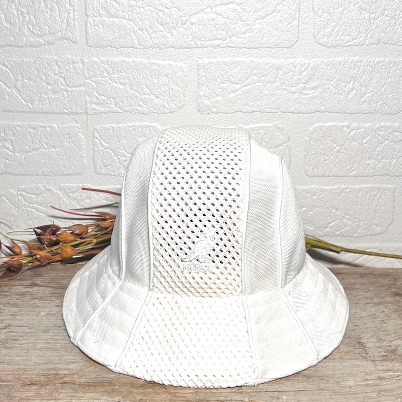 May include: A white bucket hat featuring a mesh panel design on the front and sides. The hat includes a prominent embroidered Kangol kangaroo logo on the centre front panel. The construction consists of alternating solid and mesh fabric sections.