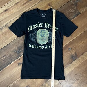 以下が含まれることがあります： 黒いTシャツ。白の筆記体で「Master Brewer Guinness & Co.」と書かれています。グラフィックには肖像画と数字の「1759」が含まれています。シャツの測定には定規が使用されています。