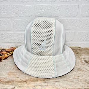 May include: A light grey bucket hat featuring a central vertical mesh panel and solid side panels. The Kangol kangaroo logo is embroidered in the centre of the mesh section. The hat has a wide, stitched brim and light blue piping accents throughout.