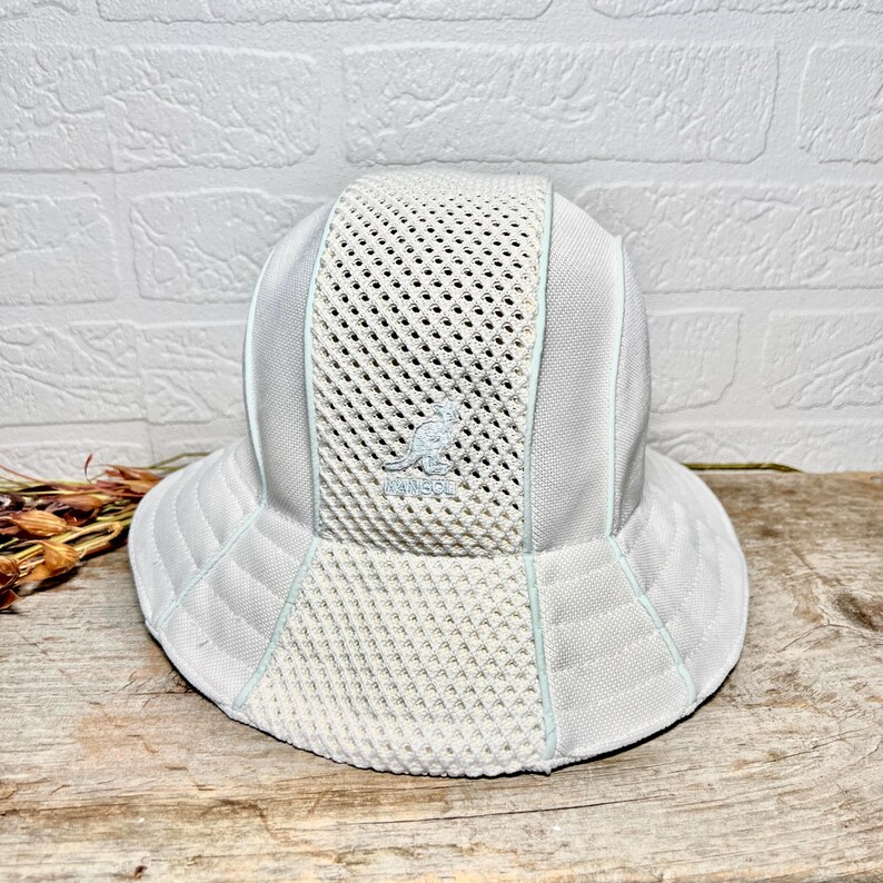 May include: A light grey bucket hat featuring a central vertical mesh panel and solid side panels. The Kangol kangaroo logo is embroidered in the centre of the mesh section. The hat has a wide, stitched brim and light blue piping accents throughout.